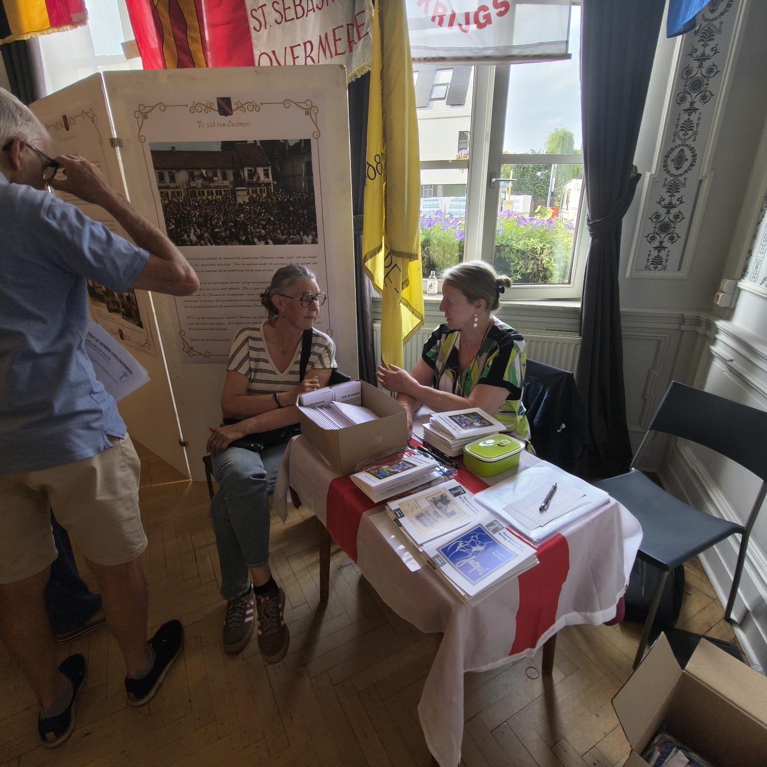 Drie mensen praten bij een tafel met boeken en documenten, binnen een ruimte met vlaggen en posters.